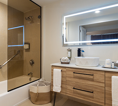 a bathroom image with a wooden vanity an led mirror a laundry basket and a glass shower