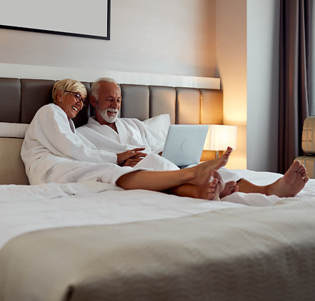 older man and woman couple looking at a laptop in bed at a hotel with hotel robes on