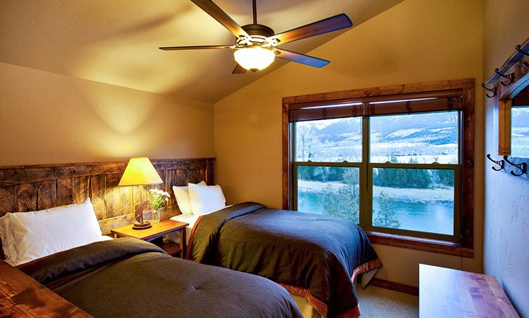 Two twin beds with a reclaimed wood headboard, nightstand with a lamp, ceiling fan and a large window.