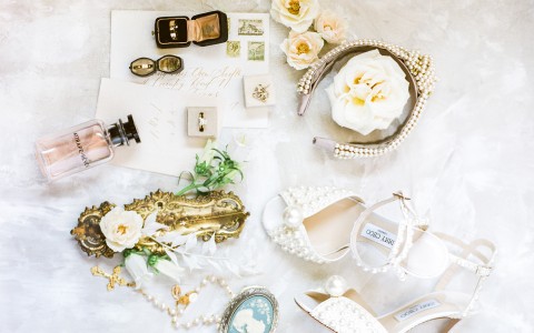 flat lay image of jimmy choo shoes, white roses, rings and other bridal accessories on a marble floor 