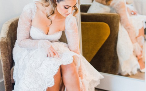 bride sitting on chair inside guest room putting on her white pearled heels, getting ready to step out for the ceremony